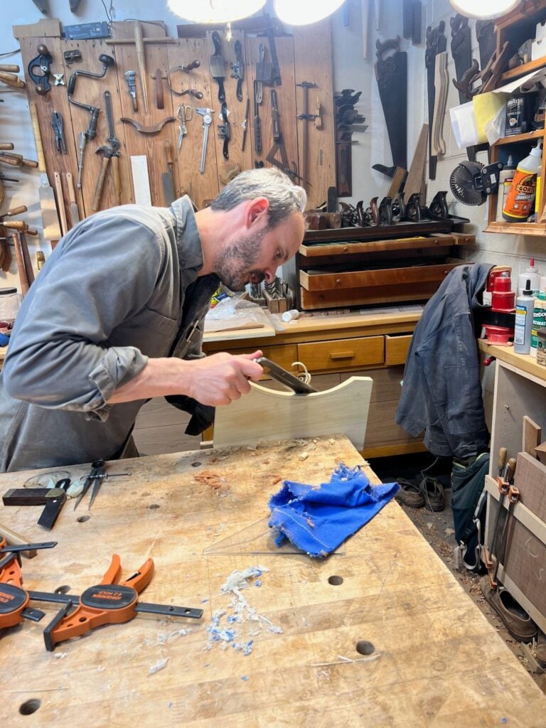 Furniture maker Evan Berding hand–sands a curved wooden component in his Durham studio, shaping a custom piece with traditional woodworking tools.