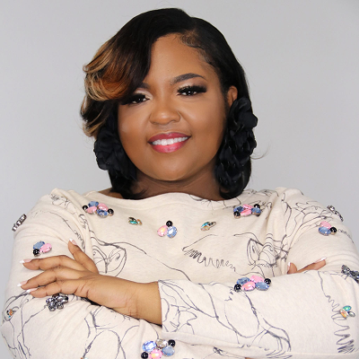 Portrait of gospel singer and preacher Jalessa Cade, smiling with arms crossed, wearing a light-colored patterned top against a neutral background.